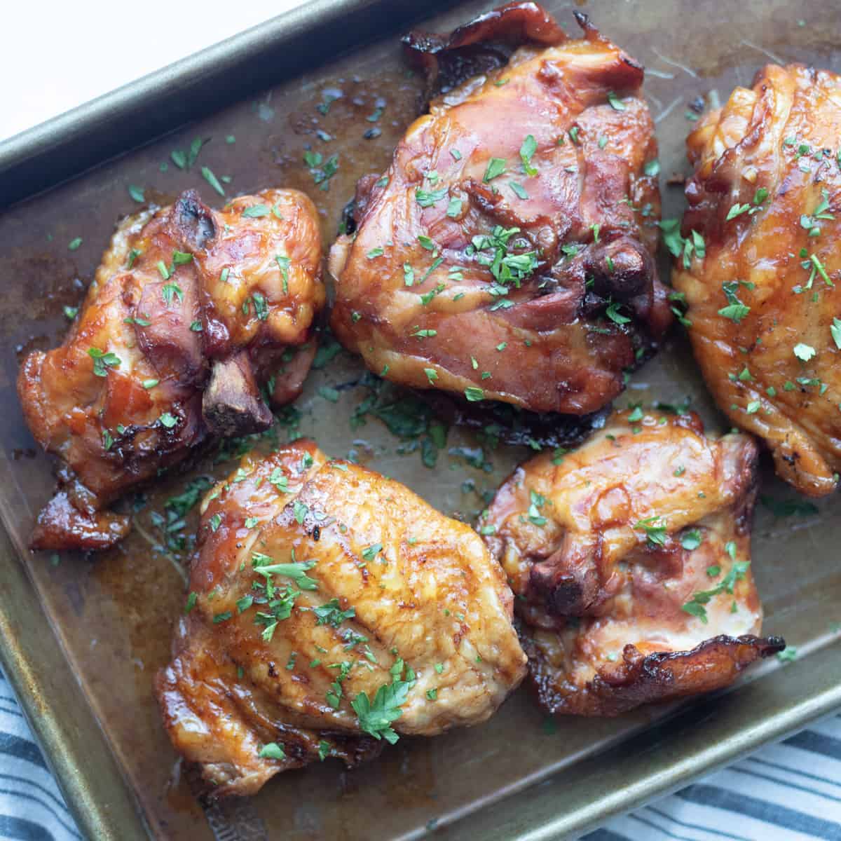 Cooked chicken thighs on a sheet pan. They're dark brown-ish orange in color and topped with chopped green parsley. 