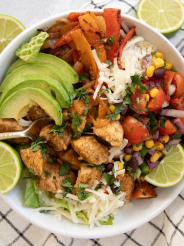 A chicken fajita taco salad in a bowl. Lettuce, chicken, peppers and onions, a tomato and bean salsa, and avocado are visible.