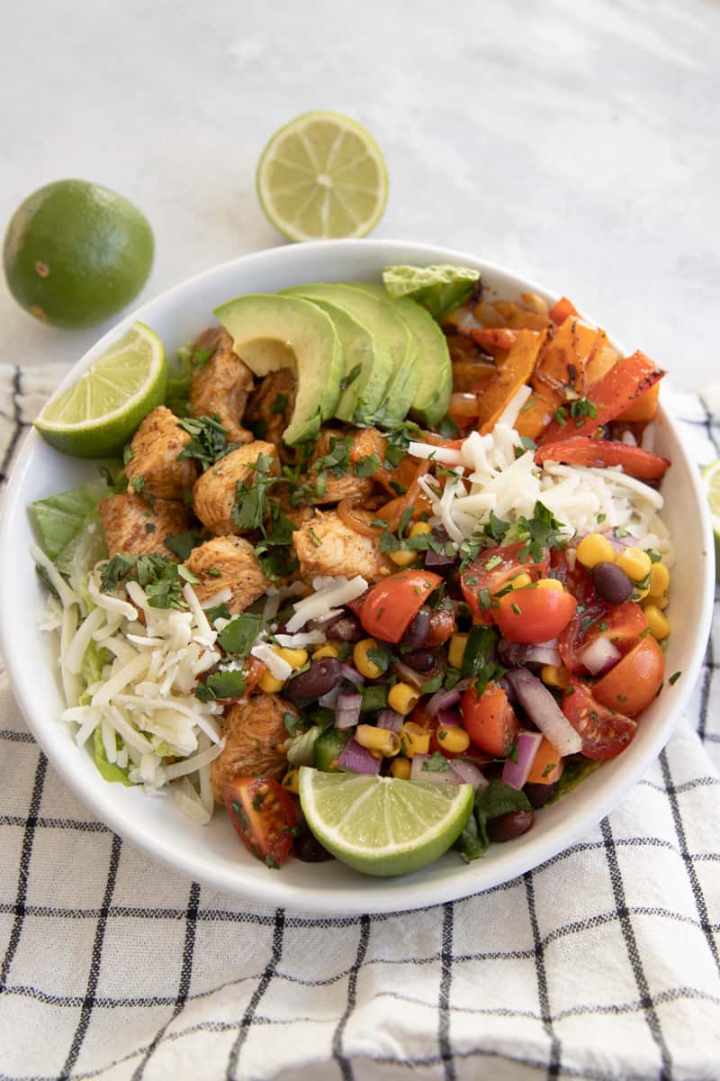 A chicken fajita taco salad in a bowl with lettuce, chicken, peppers and onions, a tomato and bean salsa, and avocado.