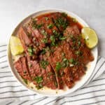 Sliced flank steak on a white plate. It's garnished with chopped cilantro and slices of limes.
