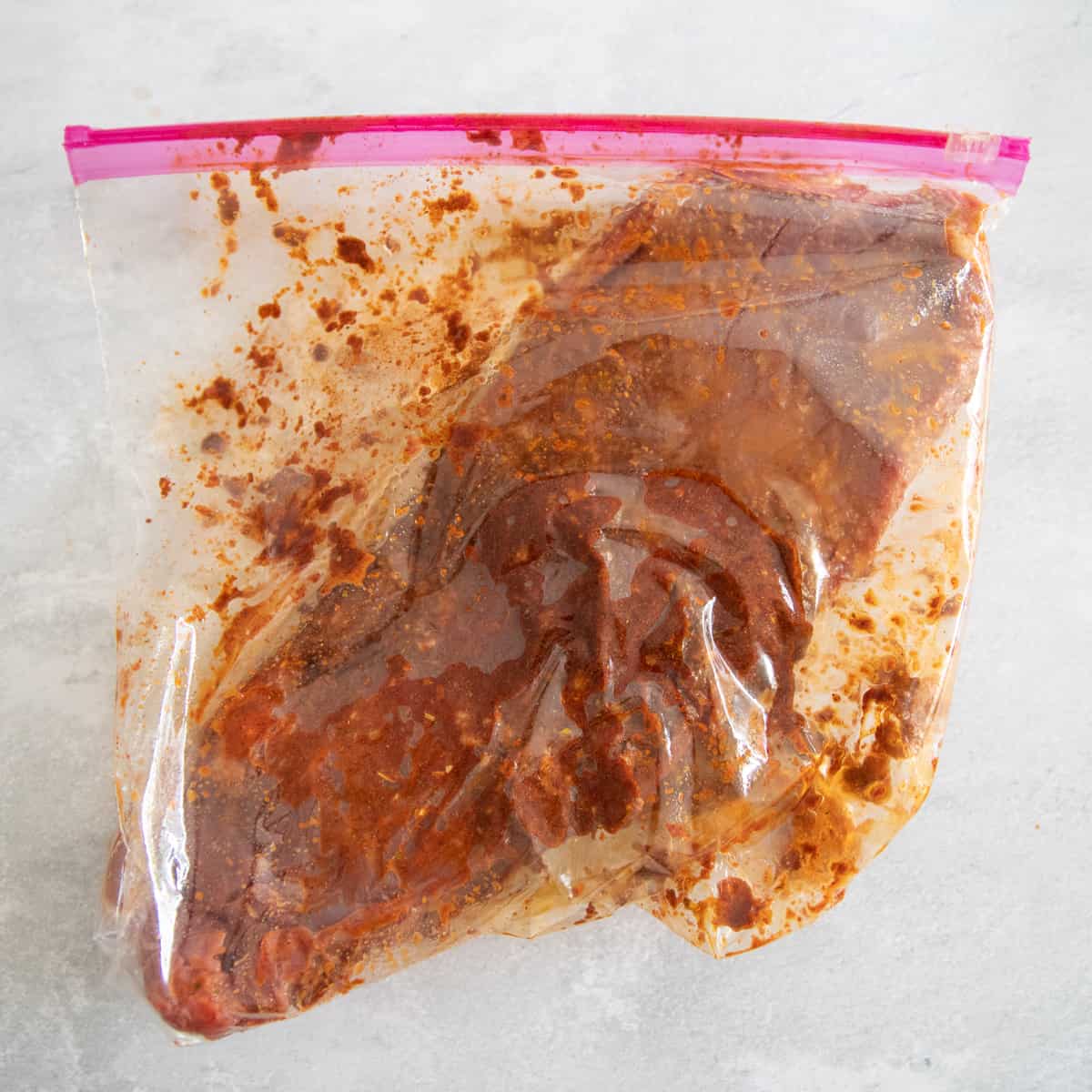 Flank steak in a Ziplock baggie with taco seasoning, oil, and lime juice. It is red-orange in color, 