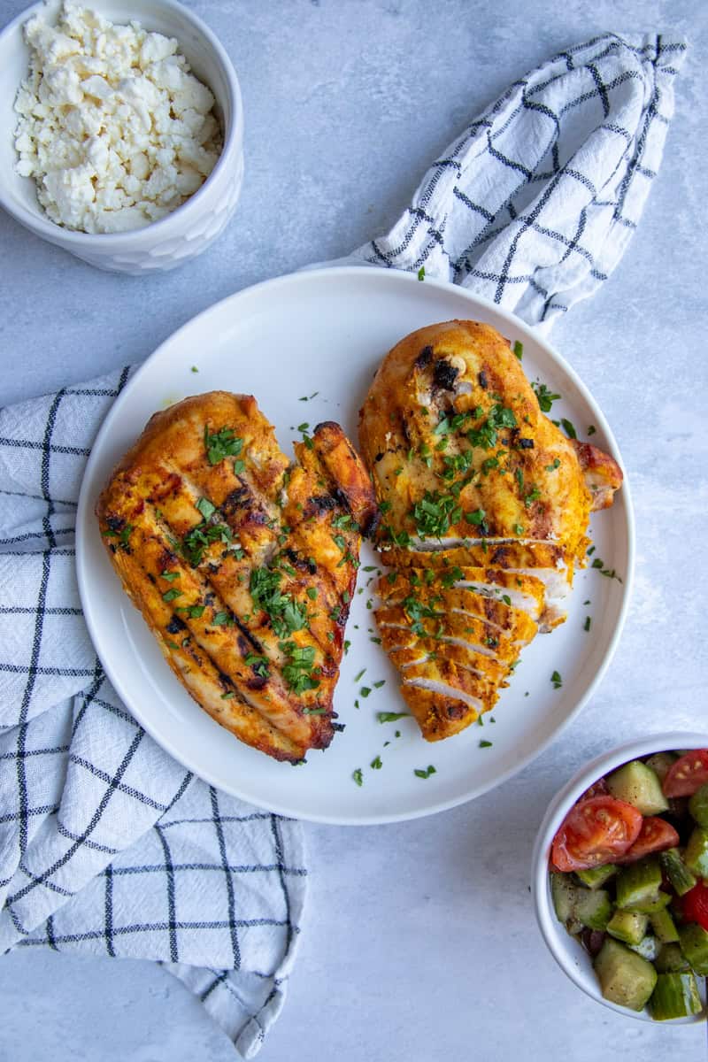Two Traeger grilled chicken breasts on a plate. They have visible grill marks, are orange-yellow in color, and are topped with green herbs.