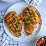 Two Traeger grilled chicken breasts on a plate. They have visible grill marks, are orange-yellow in color, and are topped with green herbs.