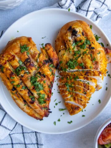 Two Traeger grilled chicken breasts on a plate. They have visible grill marks, are orange-yellow in color, and are topped with green herbs.
