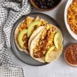 A plate of two flounder tacos topped with salad, avocado and salsa surrounded by two bowls filled with black beans and orange Mexican rice, respectively.