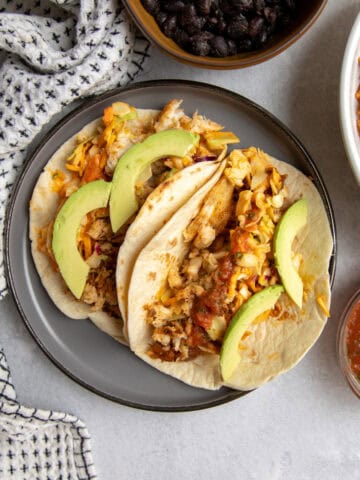 A plate of two flounder tacos topped with salad, avocado and salsa surrounded by two bowls filled with black beans and orange Mexican rice, respectively.