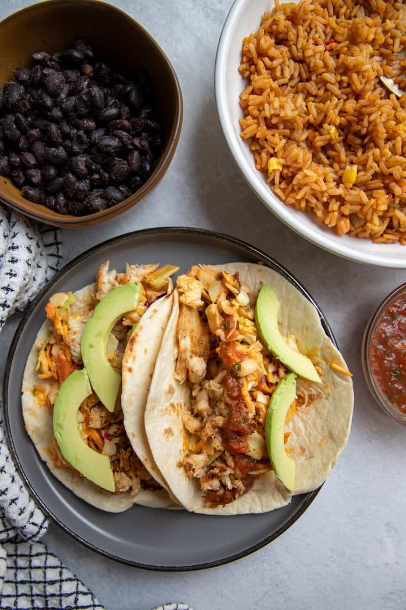A plate of two flounder tacos topped with salad, avocado and salsa surrounded by two bowls filled with black beans and orange Mexican rice, respectively.