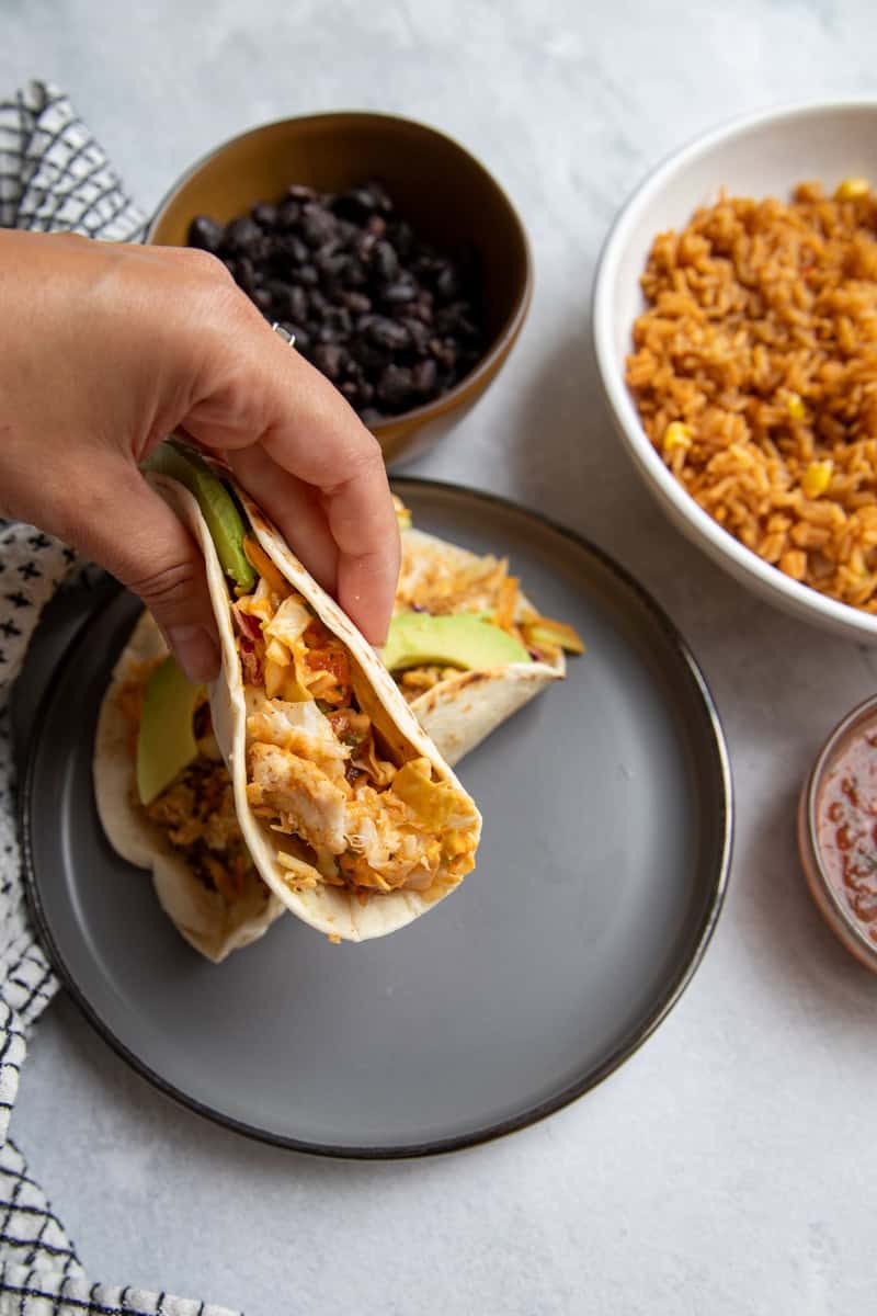 A hand holding a flounder taco topped with salad, avocado and salsa.