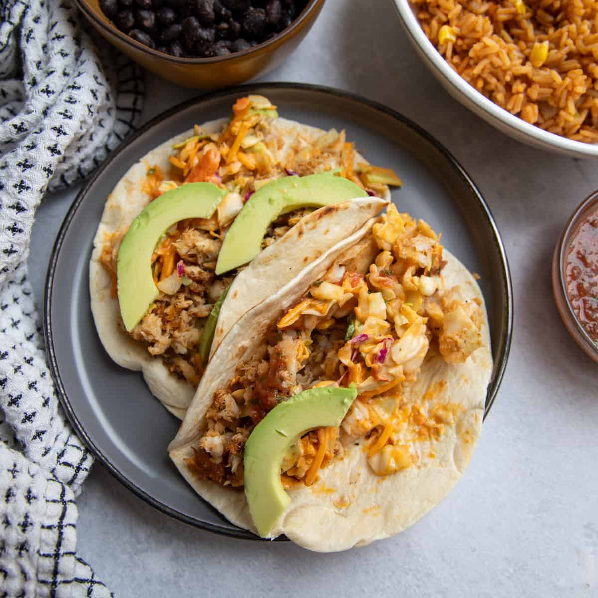 A plate of two flounder tacos topped with salad, avocado and salsa surrounded by two bowls filled with black beans and orange Mexican rice, respectively.