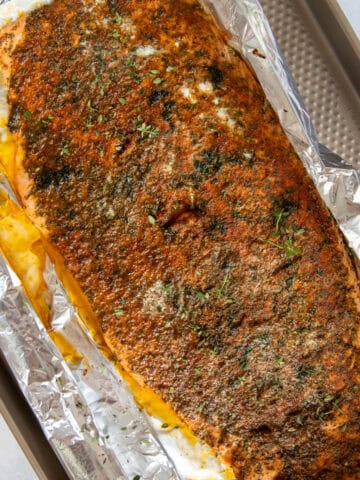 A whole cooked salmon on top of an aluminum foil-coated pan. The salmon is topped with spices and fresh dill.