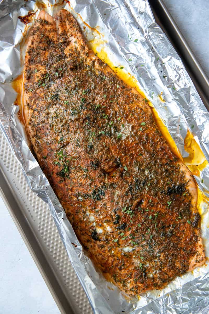 A whole cooked salmon on top of an aluminum foil-coated pan. The salmon is topped with spices and fresh dill. 
