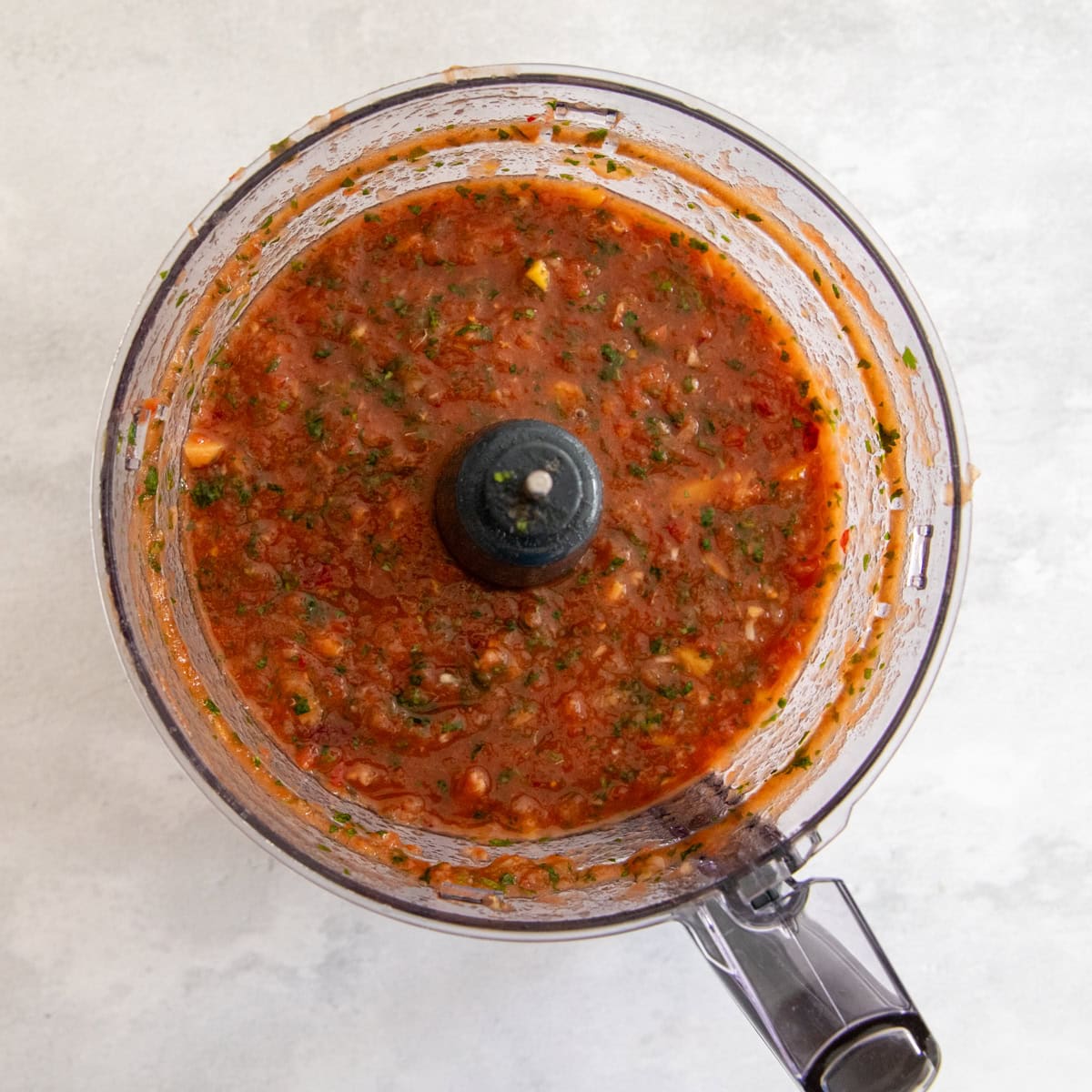 Red salsa in a food processor. It is smooth with very small chunks of tomatoes, peach, mango, and cilantro.