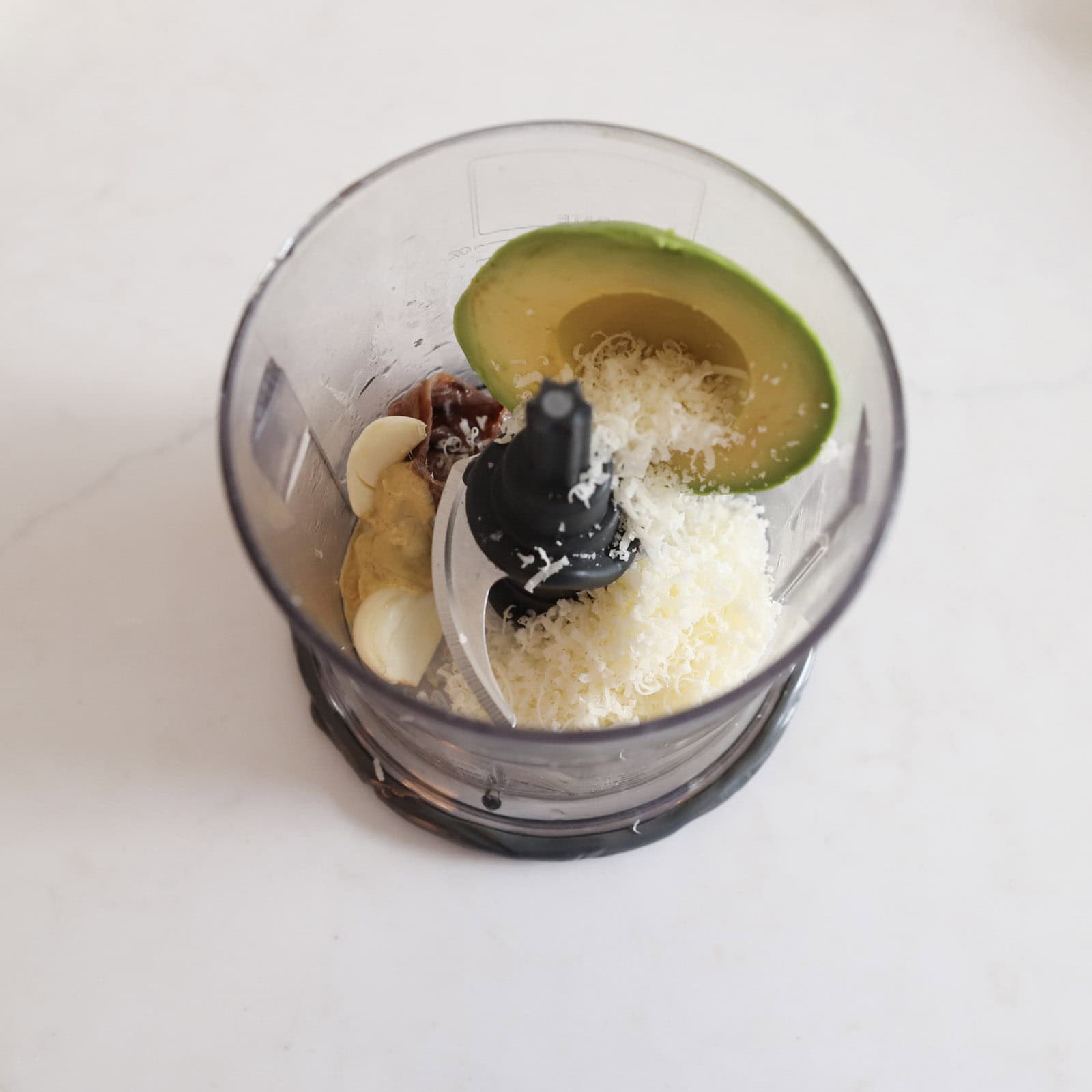 Whole avocado Caesar dressing ingredients—avocado, anchovies, Greek yogurt, mayo, parmesan, garlic, Dijon, and lime—sitting in a food processor bowl before blending.
