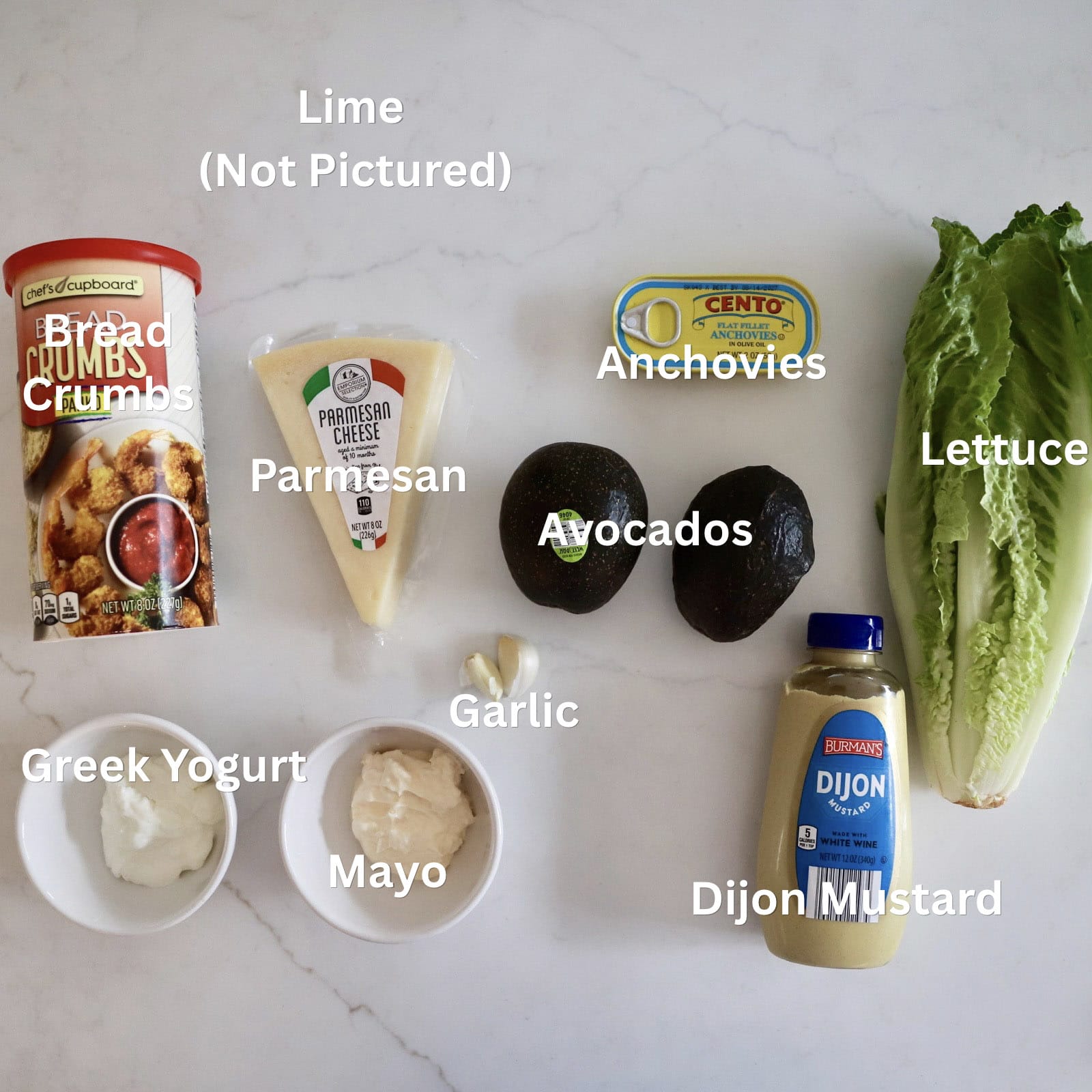 All the ingredients for avocado Caesar salad arranged on a countertop, including romaine, avocado, parmesan, anchovies, Greek yogurt, mayo, lime, garlic, Dijon, butter, and panko.