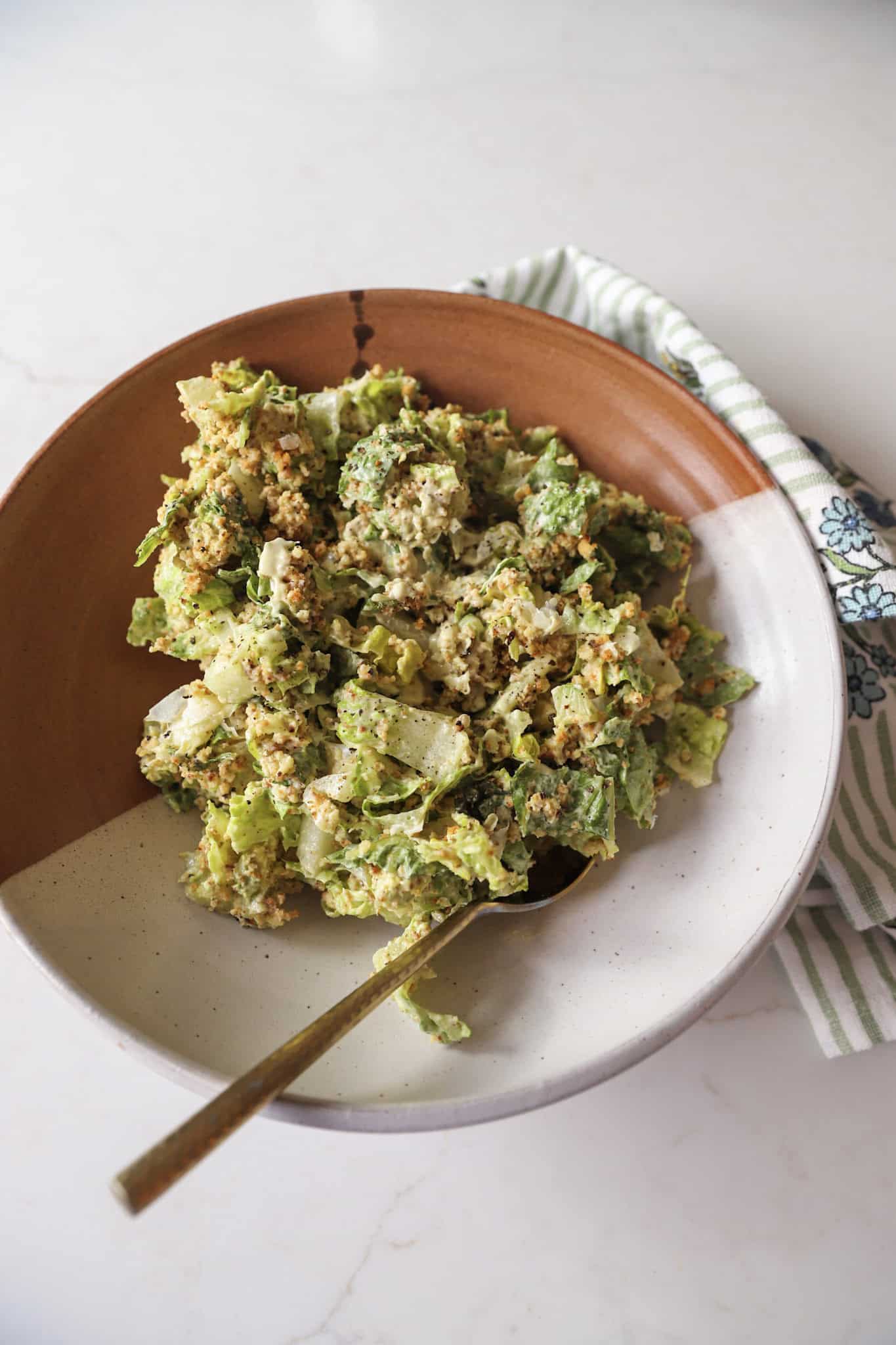 A serving bowl filled with avocado Caesar salad topped with toasted panko and parmesan, with a kitchen towel beside it.
