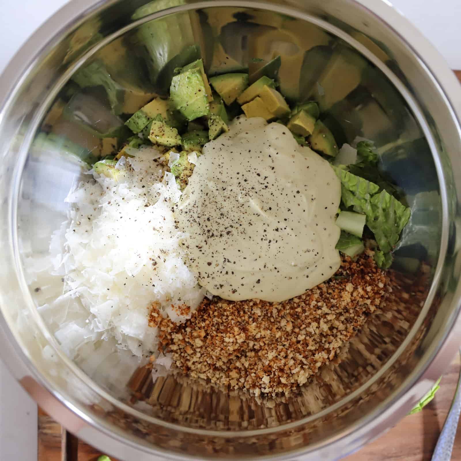 All the avocado Caesar salad ingredients—romaine, avocado, parmesan, toasted panko, freshly ground pepper, and avocado caesar dressing—in a mixing bowl, ready to be mixed together