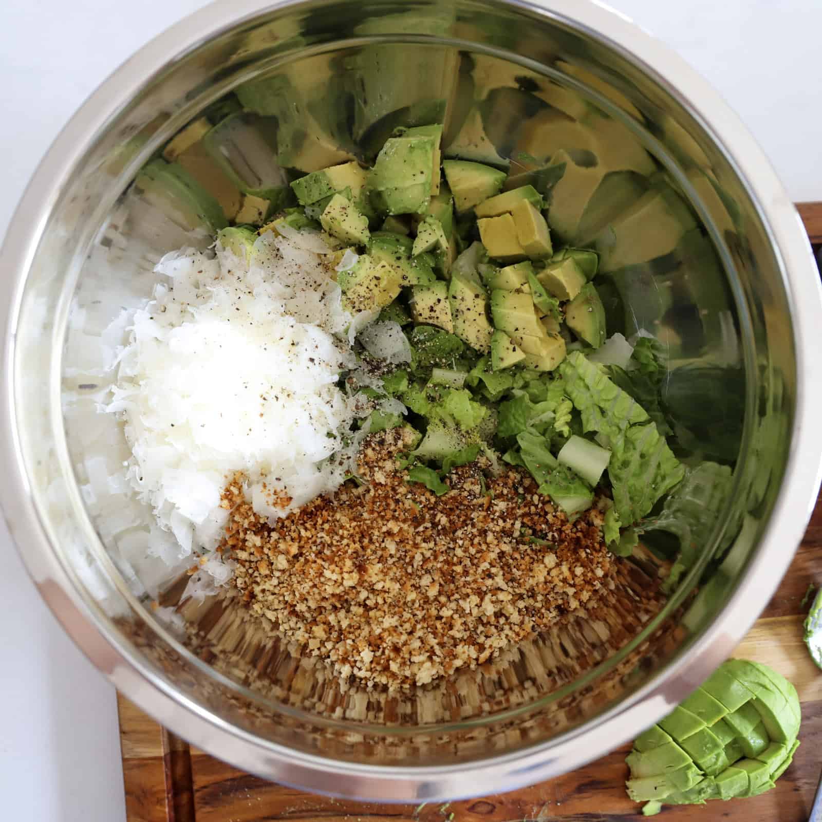 All the avocado Caesar salad ingredients—romaine, avocado, parmesan, toasted panko, and freshly ground pepper—in a mixing bowl, ready to be tossed with the dressing.